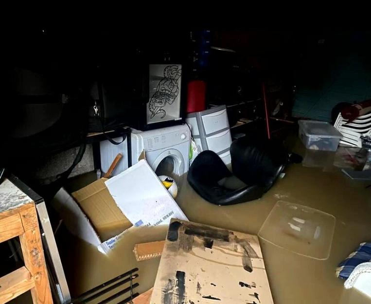 A room flooded with muddy water, containing a washing machine, boxes, and other damaged belongings.