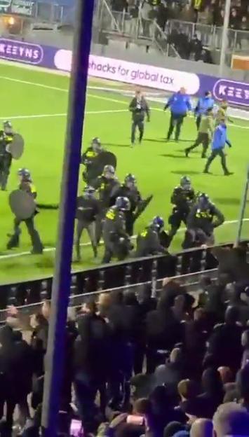 Riot police in formation on a football pitch, with spectators in the foreground.