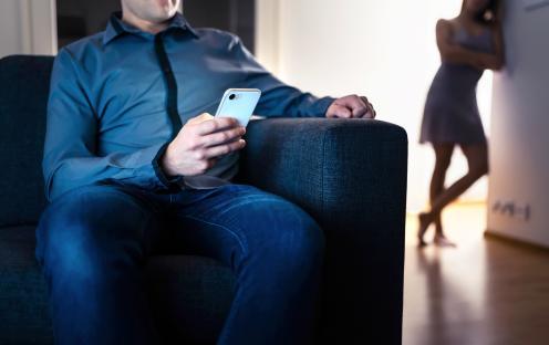 A man sits on a couch looking at his phone while a woman stands in the background.