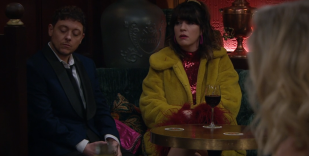 Screenshot of two people sitting at a table in a bar, a woman in a yellow coat and a man in a suit.