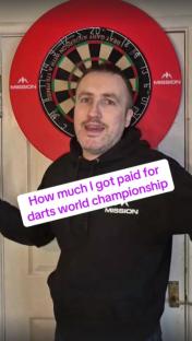 Man in front of a dartboard discussing his darts world championship earnings.