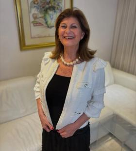 A woman wearing a white jacket and pearl necklace smiles at the camera.