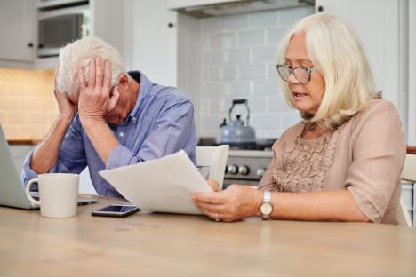 Senior couple reviewing financial documents at their kitchen table, looking distressed.