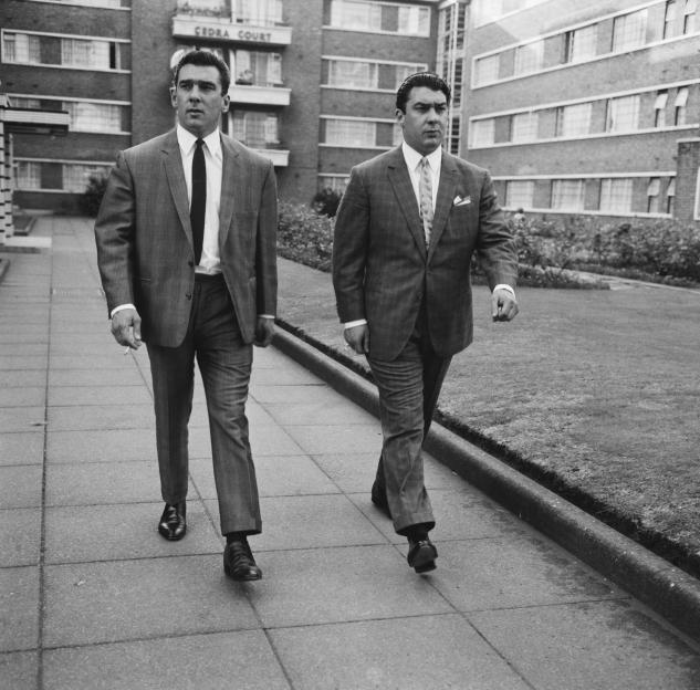Black and white photo of twin brothers, Ronnie and Reggie Kray, walking down a sidewalk.