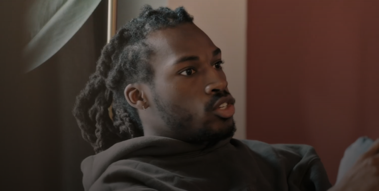 A man with dreadlocks wearing a hoodie looks right and speaks.