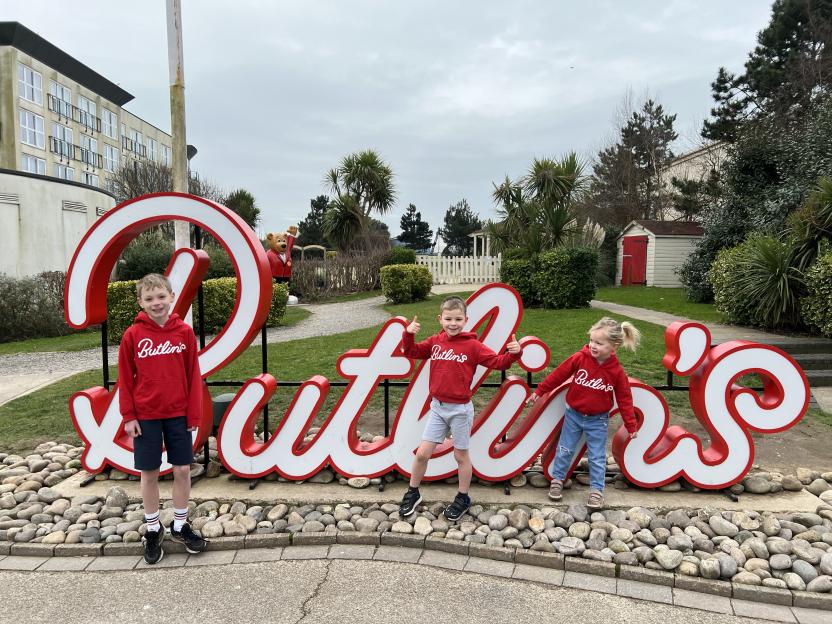 Three children in red Butlin's sweatshirts stand in front of a large Butlin's sign.