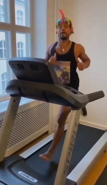 A man wearing a black tank top and a colorful mohawk headpiece runs barefoot on a treadmill.