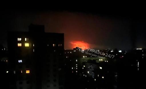 Nighttime view of a city with a large fire or explosion in the distance.