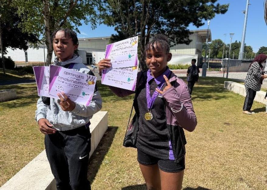 Two girls holding certificates and a medal outdoors.