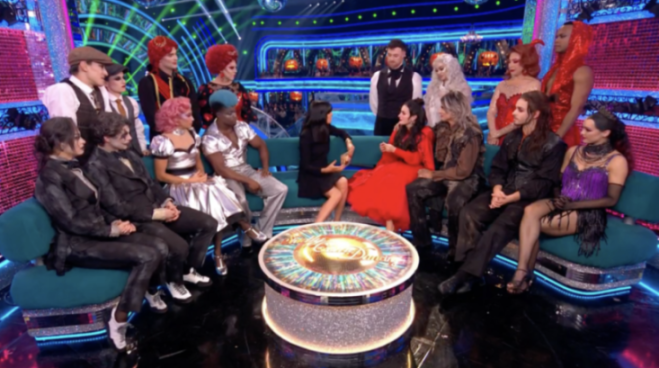Contestants in various costumes sit on green couches around a sparkly table with the "Strictly Come Dancing" logo.