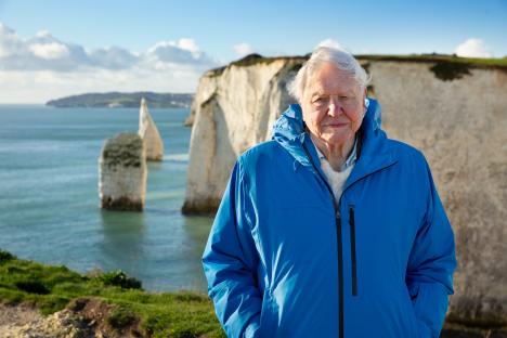 Sir David Attenborough standing outdoors near a body of water and chalk cliffs.