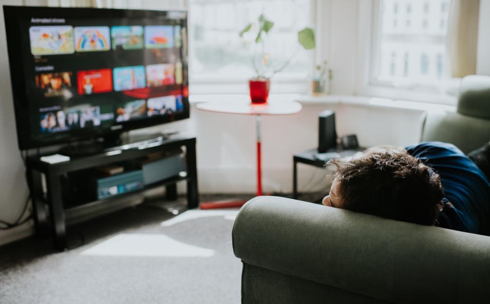 A man lies on a sofa, staring at a large, flatscreen TV displaying a choice of shows on a streaming platform