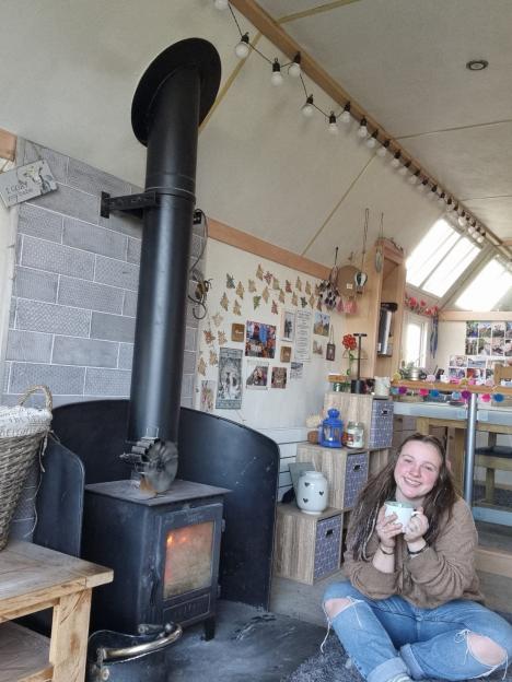 Woman sitting in her off-grid static caravan by a wood-burning stove.