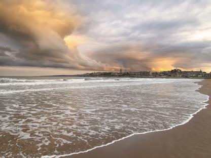 Sunset over a beach with waves and a town in the distance.