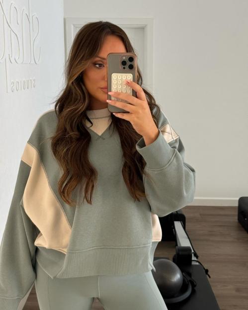 Woman with long brown hair wearing a two-tone sweatshirt and leggings, taking a mirror selfie with a phone.