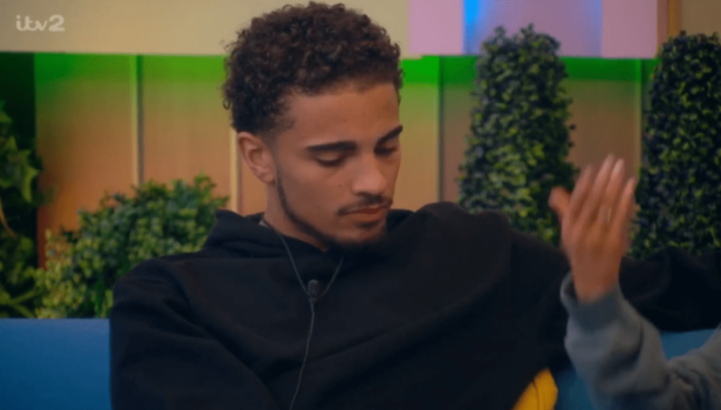 A man in a black hoodie with a mic attached to his shirt sits on a blue couch, looking down, while a woman's hand is visible next to him.