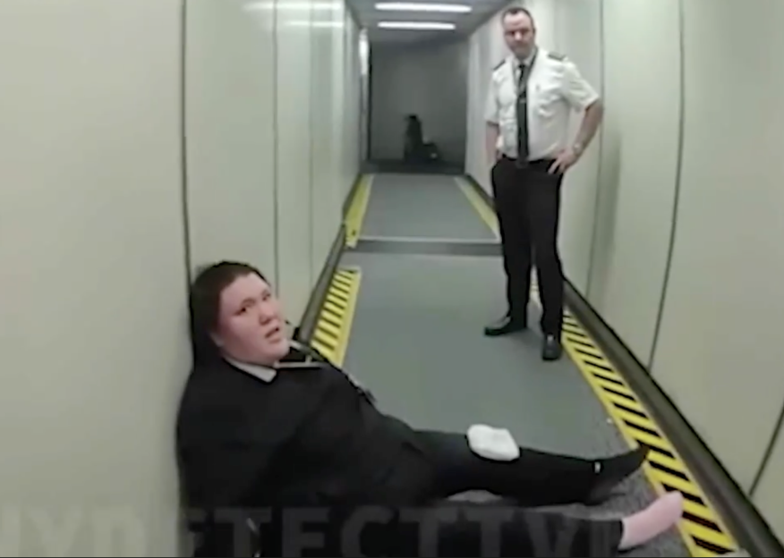 A person in black clothing is sitting on the floor of a jet bridge, while a pilot stands over them.