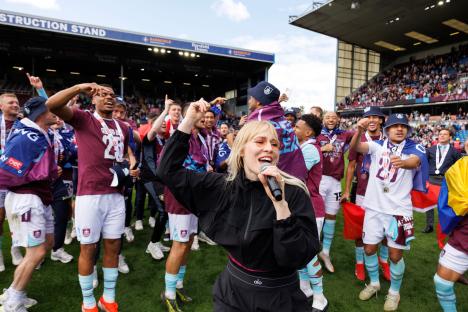 Natasha Bedingfield singing on a football pitch with Burnley players celebrating.