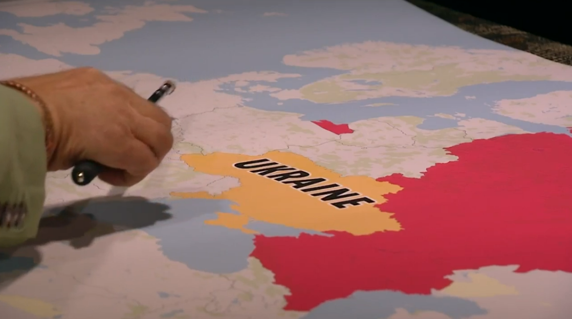 A hand with a pen pointing to a map of Ukraine colored yellow, with surrounding countries in red.