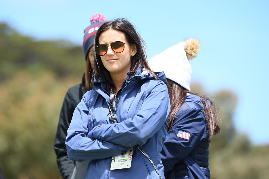 Jillian Wisniewski in sunglasses, wearing a blue jacket with arms crossed, watching a golf match.