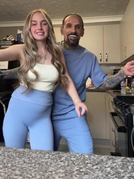 A young woman and a man in a kitchen, both smiling and wearing blue, with the woman wearing a cream top and the man a blue t-shirt and showing off tattoos on his arms.