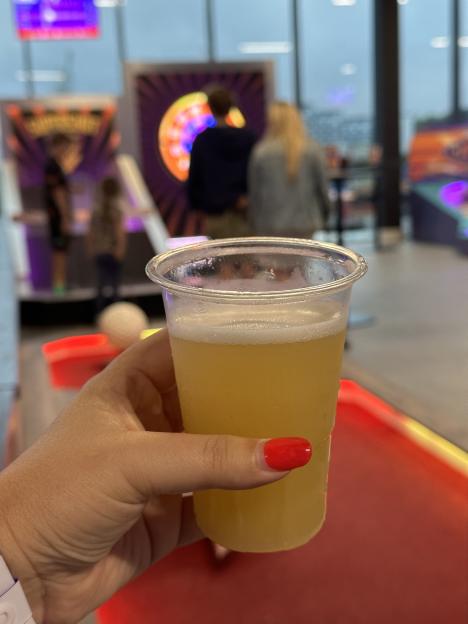 A hand holding a plastic cup of light yellow beer in an arcade.