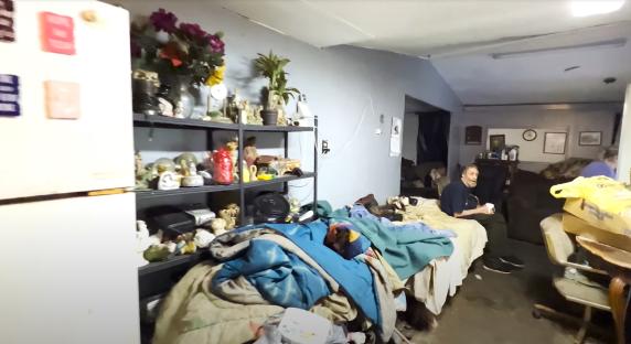 Interior of a cluttered home with a man sitting on a bed.