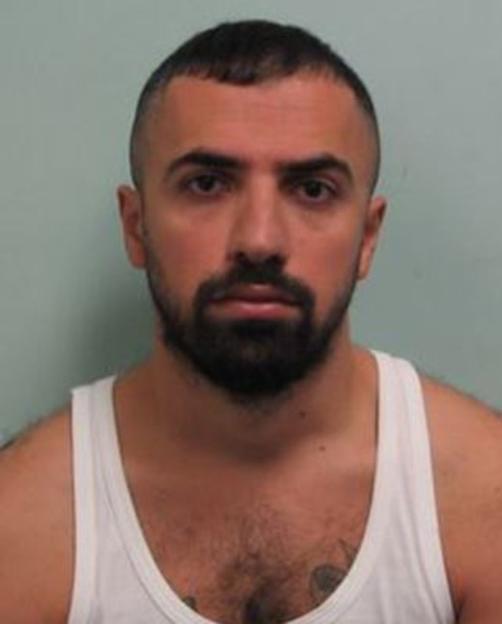 Mugshot of a man with short dark hair and a beard, wearing a white tank top.