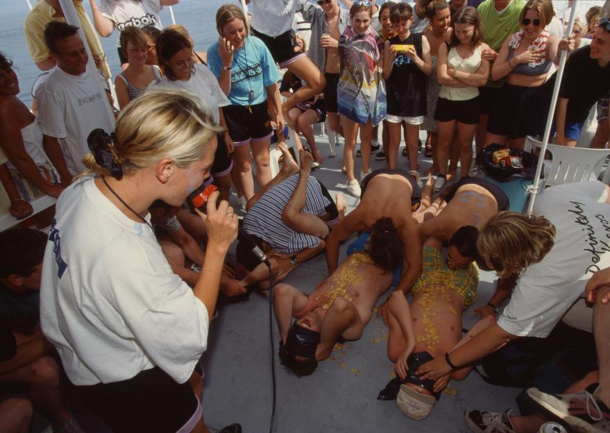A group of young adults on a boat participate in a game where people with blindfolds have potato chips placed on their chests.