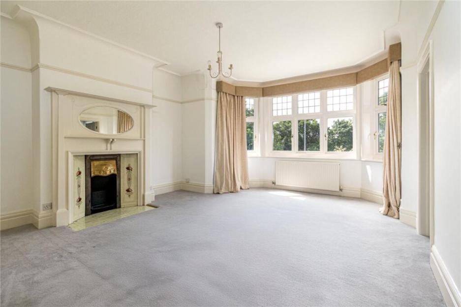 An empty living room with a large bay window overlooking trees, a fireplace with an ornate mantelpiece, and light gray carpeting.