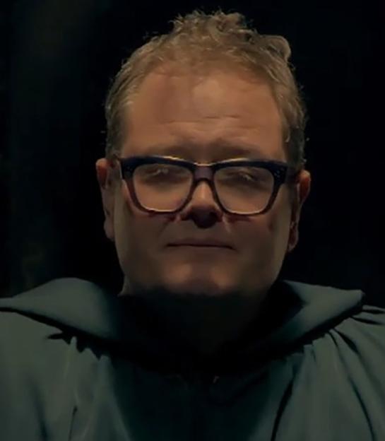 Alan Carr in a dark cloak and glasses, with two lanterns behind him.