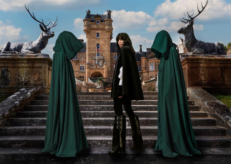 Claudia Winkleman on a stone staircase between two cloaked figures, with a castle and deer statues in the background.
