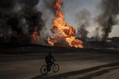 A person on a bicycle rides past burning buildings.