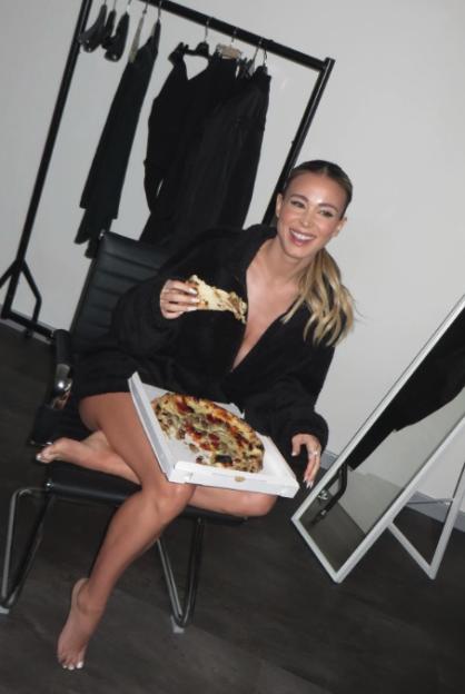 A woman in a black robe eats pizza while sitting in a black chair, with a clothing rack behind her.