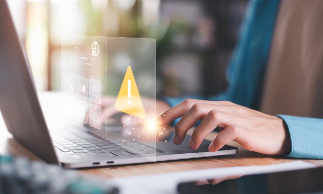 A person working on a laptop with a prominent warning sign (exclamation mark in a triangle) overlaid on the screen, indicating a computer virus or system error.