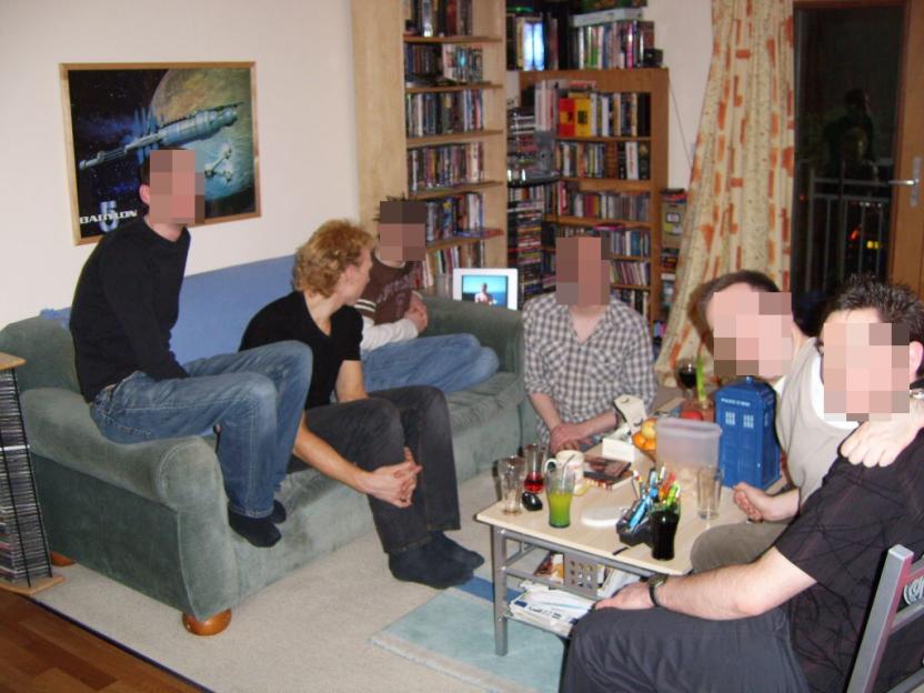 Five men gathered in a living room, with three sitting on a sofa and two at a small table, surrounded by bookshelves and a "Babylon 5" poster.