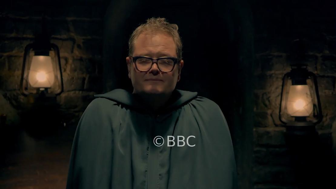 Alan Carr in a dark cloak and glasses, framed by two lanterns, with the BBC logo visible.