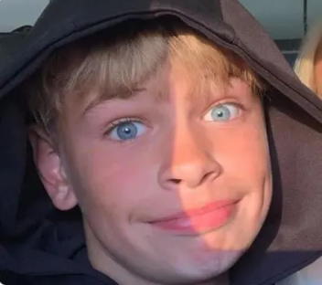 Close-up of a boy with blond hair and blue eyes, wearing a dark hoodie.