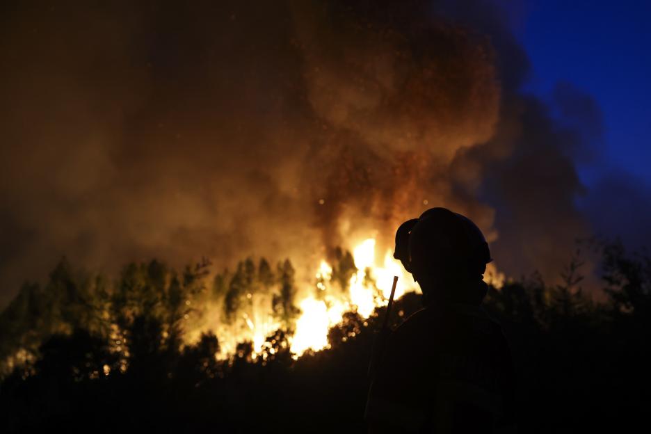 Silhouetted firefighter observing a large forest fire at night.