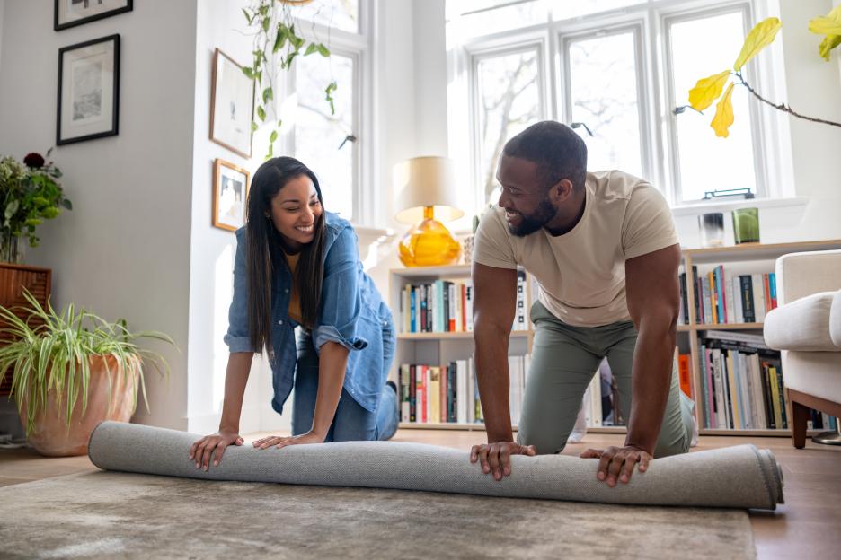 Happy African American couple unrolling a new carpet at home while smiling.