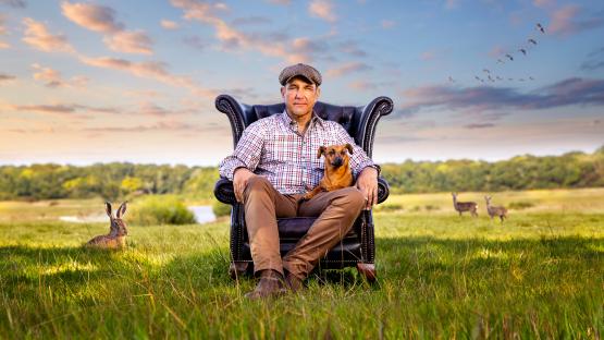 Man sitting in a chair outdoors with a small dog, rabbits, and deer in the background.