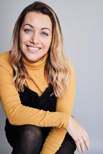 Headshot of a smiling woman with blonde hair wearing a mustard-colored turtleneck and black dress.