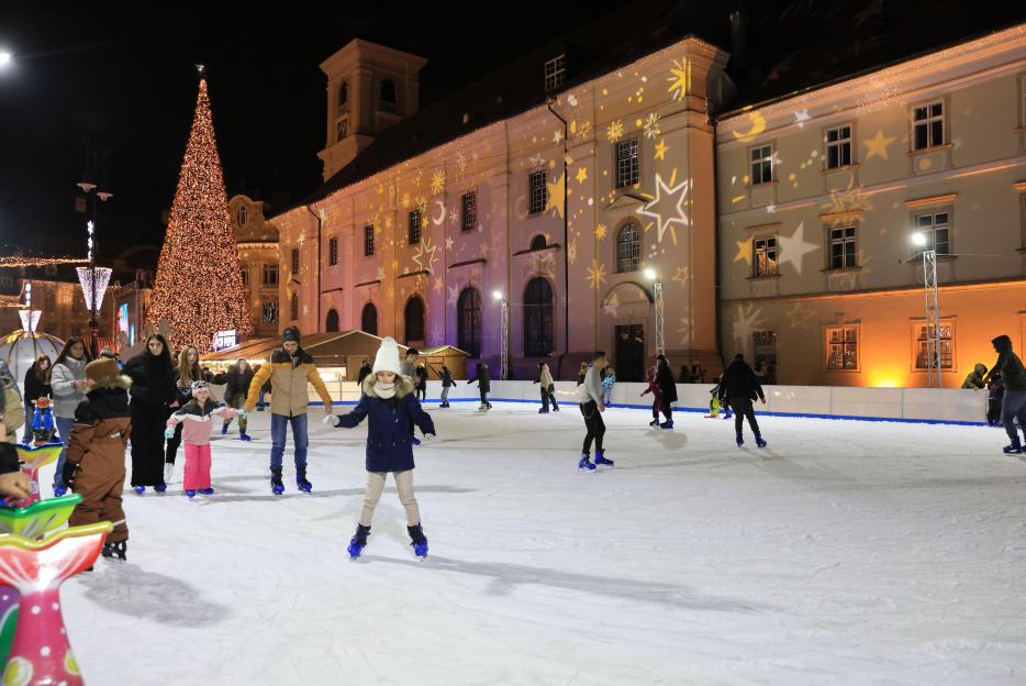 People ice skating on Piata Mare in Sibiu, Romania, with a large Christmas tree and buildings with star projections in the background.