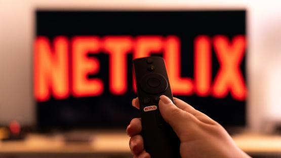 Hand holding a Netflix remote in front of a TV displaying the Netflix logo.