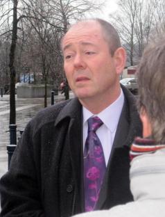 A man in a dark coat and purple tie speaks outside.
