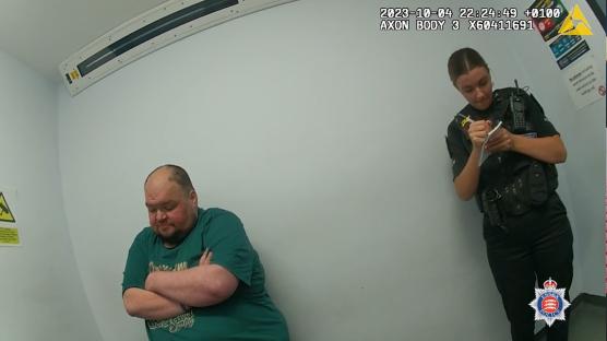Police officer taking notes while Gavin Plumb stands with arms crossed.