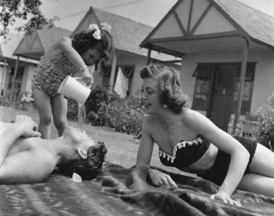 A young girl pours water on her sleeping father while their mother watches.
