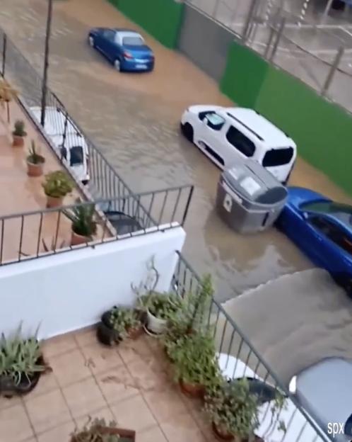 Flooded street in Ibiza with a blue sedan, a white van, and a blue sports car partially submerged in brown water.