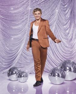 Nikita Kuzmin in a brown suit, posing against a sparkly background with disco balls.