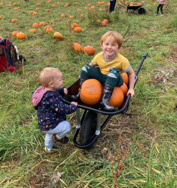 Ralph sitting in a wheelbarrow with pumpkins while his younger brother Max stands beside him in a pumpkin patch.